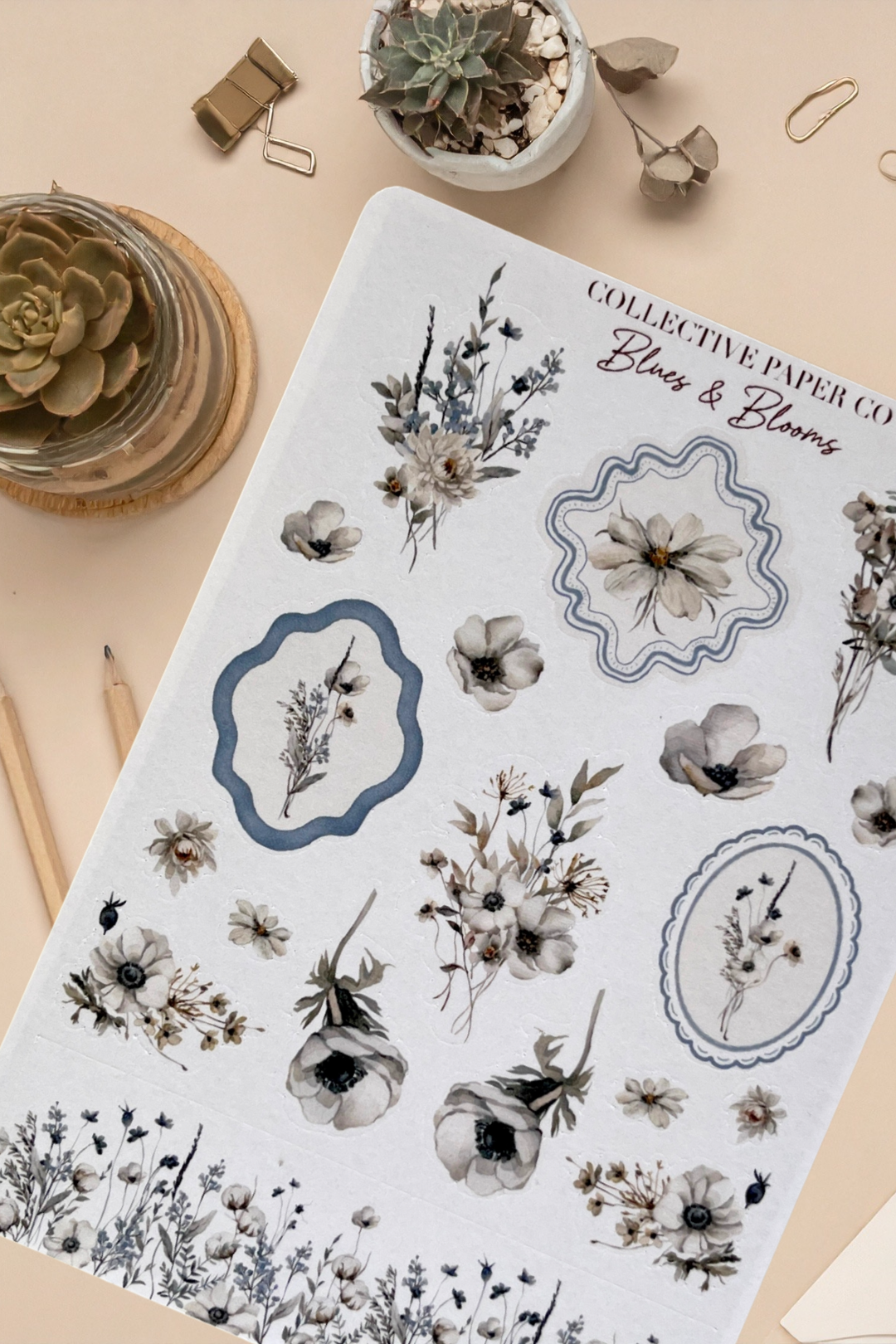 Sheet of floral stickers with decorative frames on a beige surface with plants and stationery items.