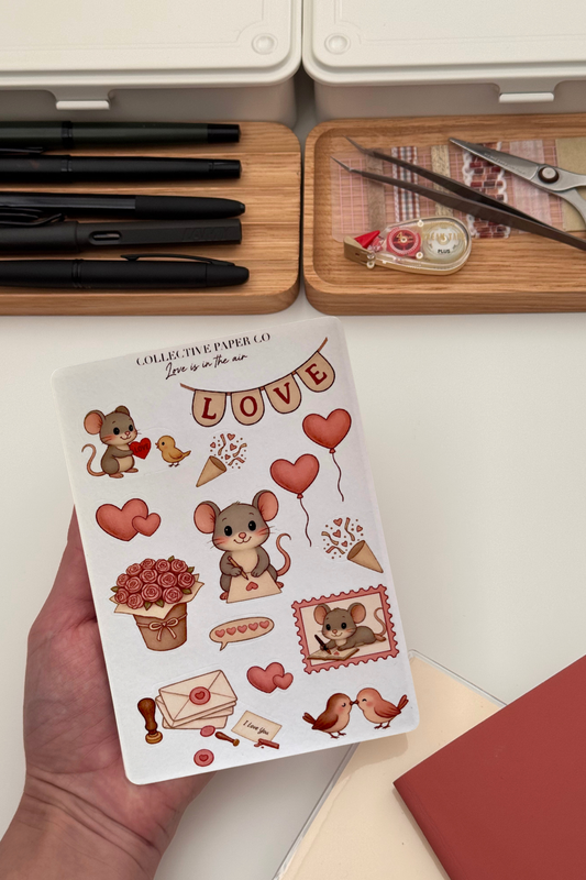 Sheet of stickers with cartoon mice and hearts held by a hand, with stationery items in the background.