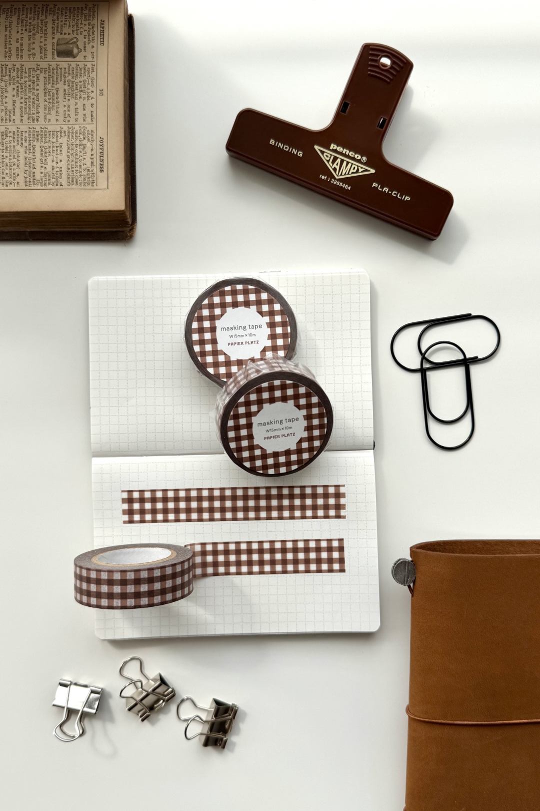 Brown gingham masking tape rolls, grid notebook with tape strips, clips, leather journal, vintage book, and red binding clip in a flat lay stationery setup.