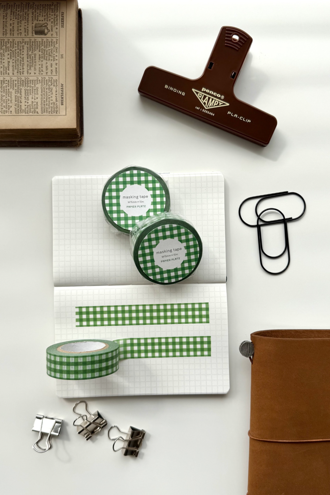 Green gingham masking tape rolls, grid notebook with tape strips, clips, leather journal, vintage book, and red binding clip in a flat lay stationery setup.