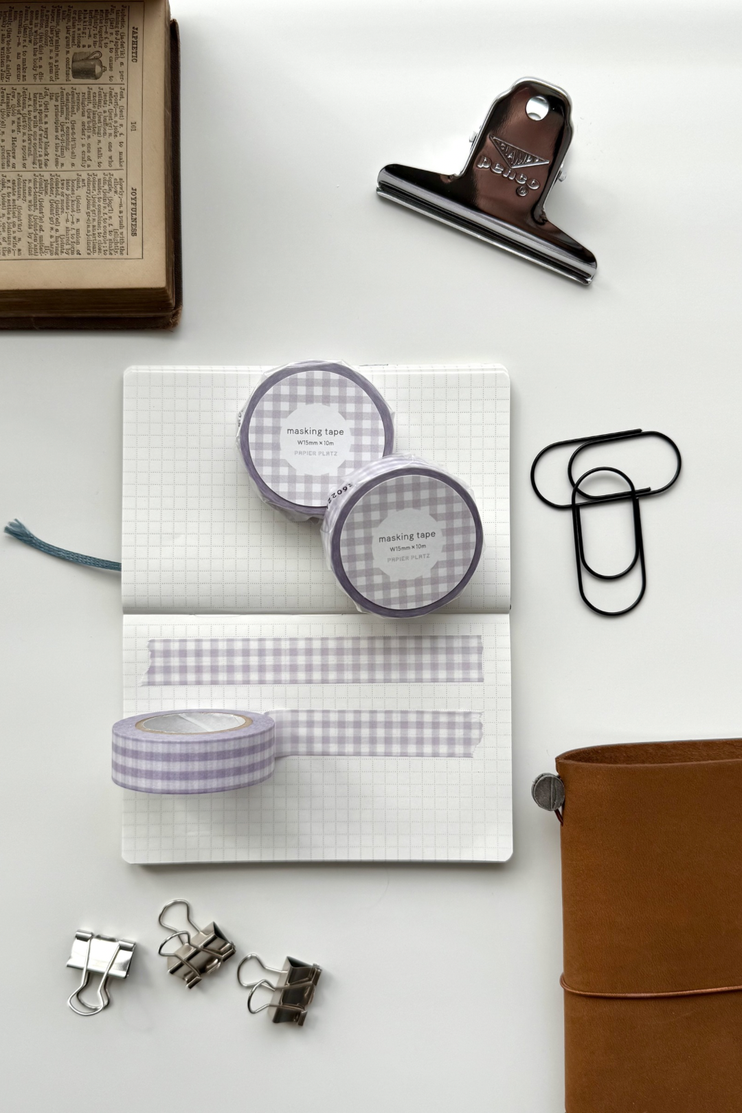 Lavender gingham masking tape rolls, grid notebook with tape strips, clips, leather journal, vintage book, and red binding clip in a flat lay stationery setup.