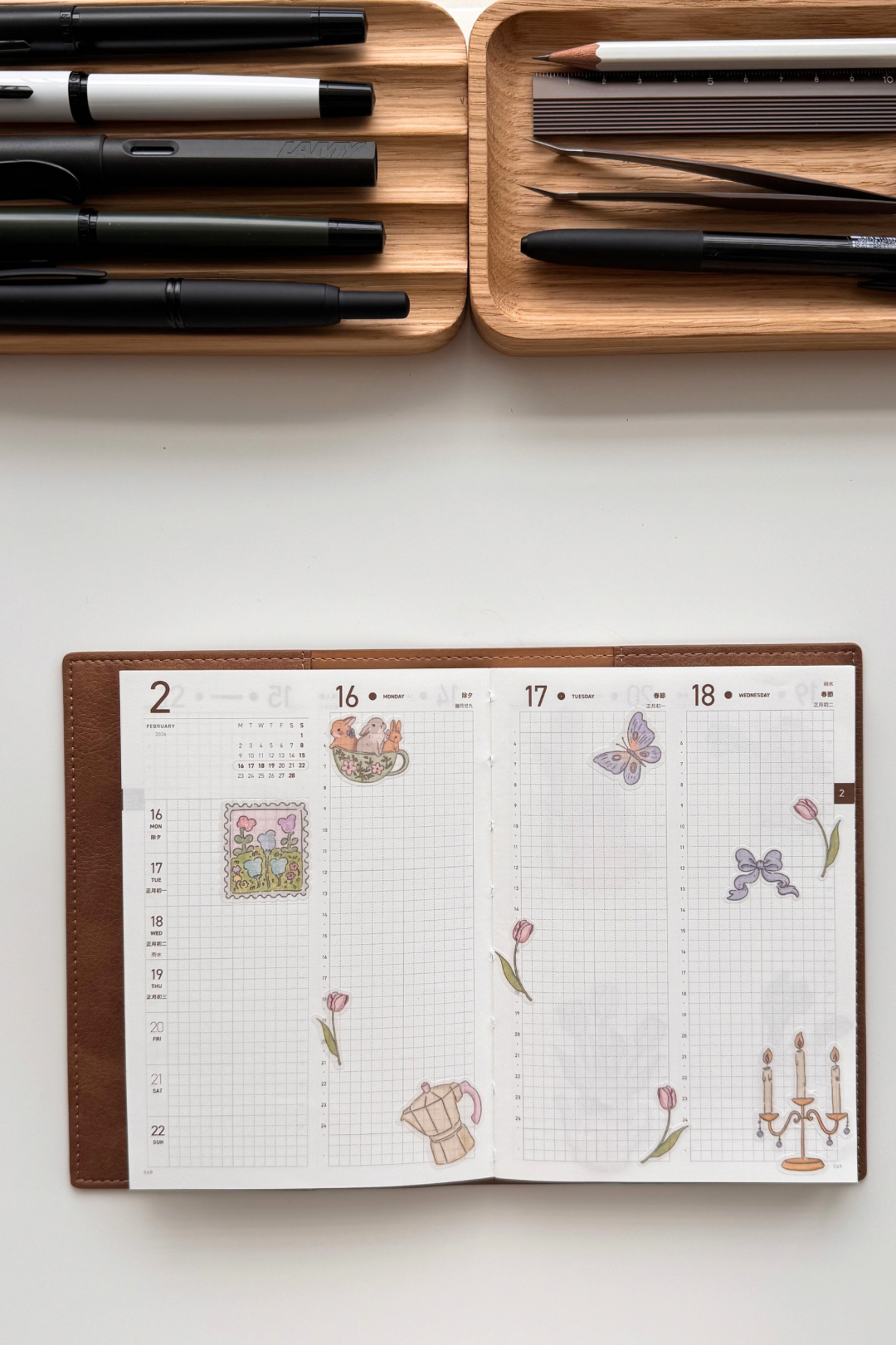Open double-page spread from a weekly journal on grid paper, decorated with pastel watercolour-style stickers including purple butterflies, pink tulips with leaves, a bunny in a teacup, a coffee pot, candelabra, postage stamp motifs and floral arrangements in soft pinks and greens.