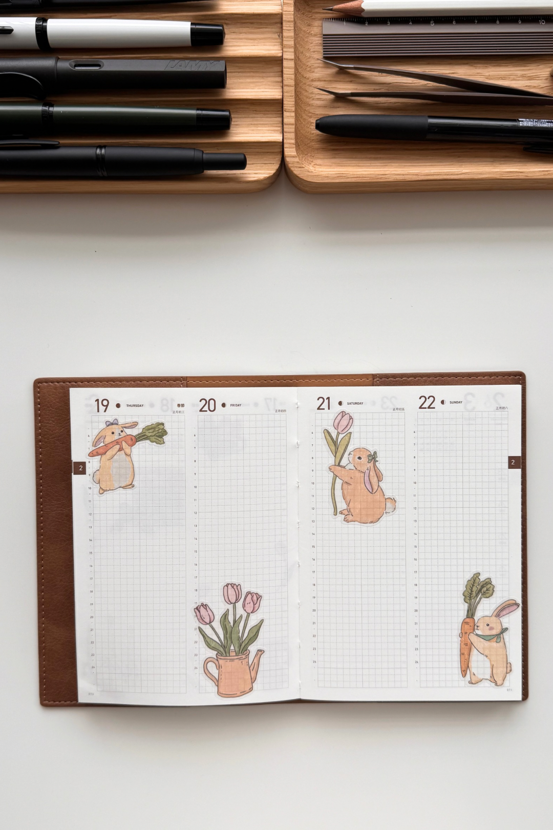 Open double-page spread from a weekly planner on faint grid paper, decorated with soft pastel watercolour illustrations of cute chubby orange bunnies holding carrots and tulips, pink tulips in a vase and with leaves, arranged playfully across the blank pages.