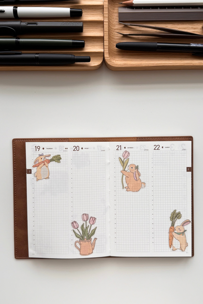 Open double-page spread from a weekly planner on faint grid paper, decorated with soft pastel watercolour illustrations of cute chubby orange bunnies holding carrots and tulips, pink tulips in a vase and with leaves, arranged playfully across the blank pages.