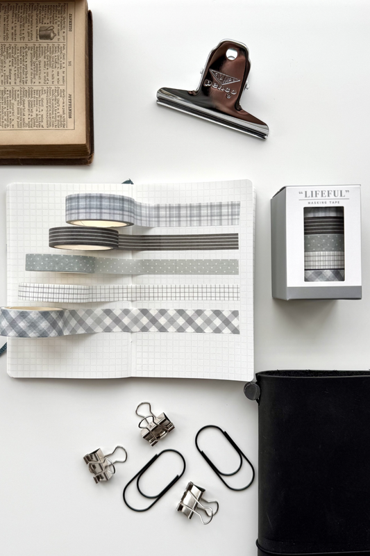 Gray and white patterned masking tapes from Lifeful on open grid notebook, with clips, black journal, vintage book, and red clip in flat lay setup.