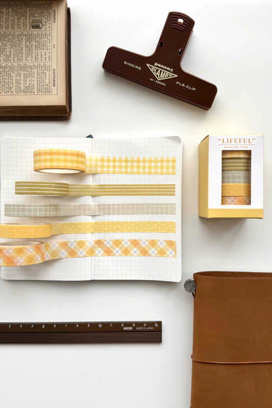 Yellow and white patterned masking tapes from Lifeful on open grid notebook, with clips, brown journal, vintage book, and red clip in flat lay setup.

