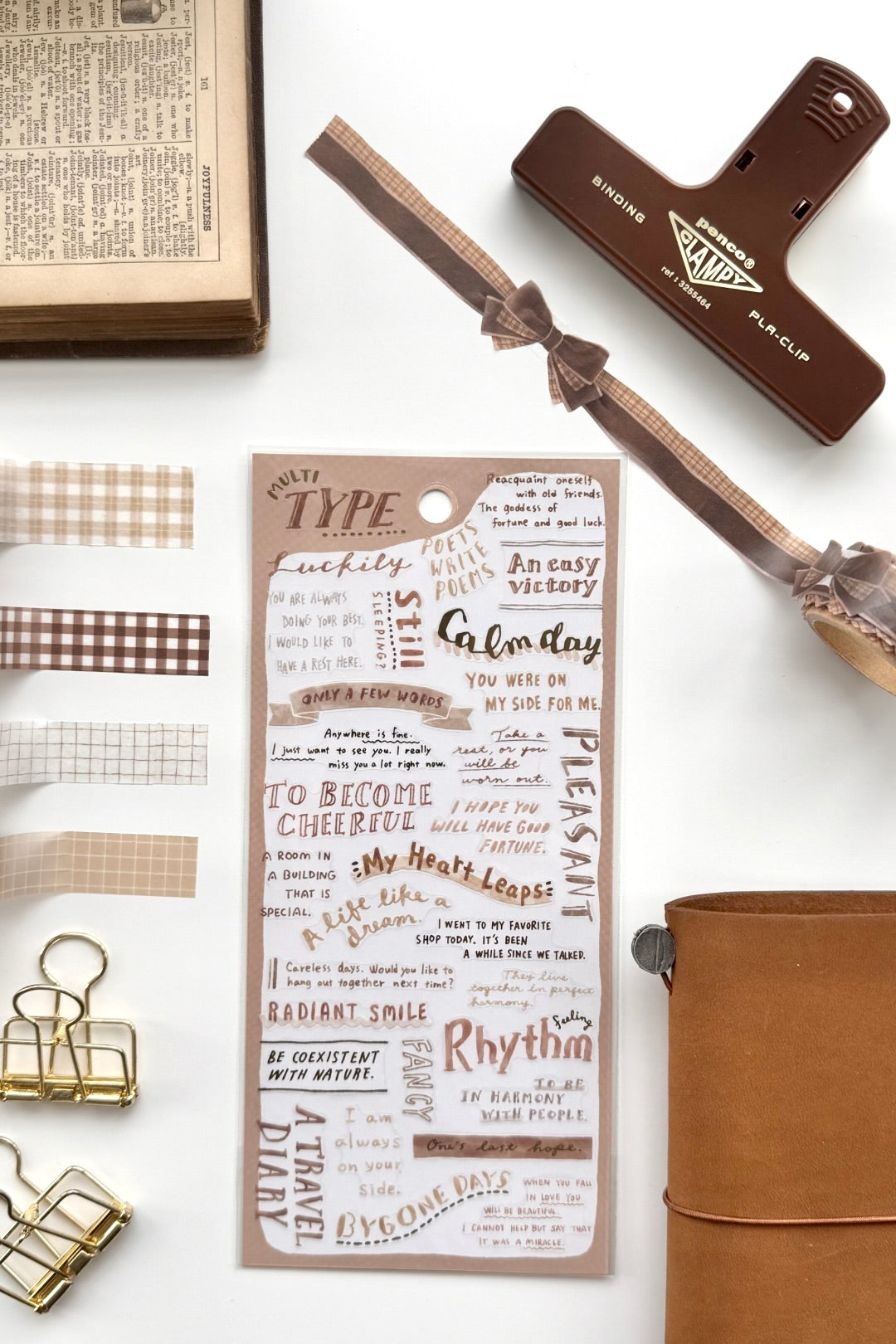 "Type Club" brown inspirational quote washi tape sheet with checkered edges, red binder clip, gold clips, book, leather journal in flat lay.