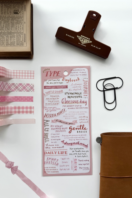 "Type" pink washi tape sheet with quotes like "Glorious Day," "Mystery," matching checkered tapes, bow, red clip, book, leather journal in flat lay.

