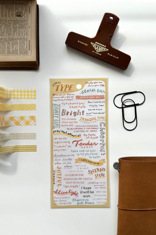 "Type" yellow washi tape sheet with quotes like "Genial Day," "Tender," matching checkered tapes, red clip, book, leather journal in flat lay.

