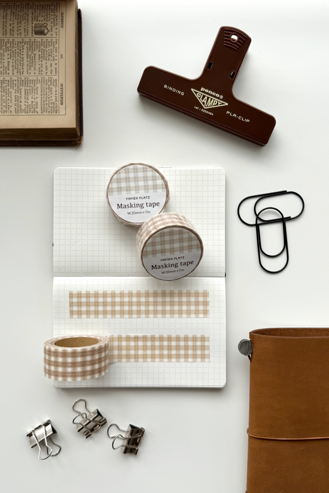 A flat lay composition features two rolls of beige gingham masking tape, an open graph paper notebook with tape applied, various metal clips and binder clips, a brown leather journal, an old book, and a red vintage stapler on a white surface. The aesthetic evokes cozy journaling or scrapbooking stationery setup