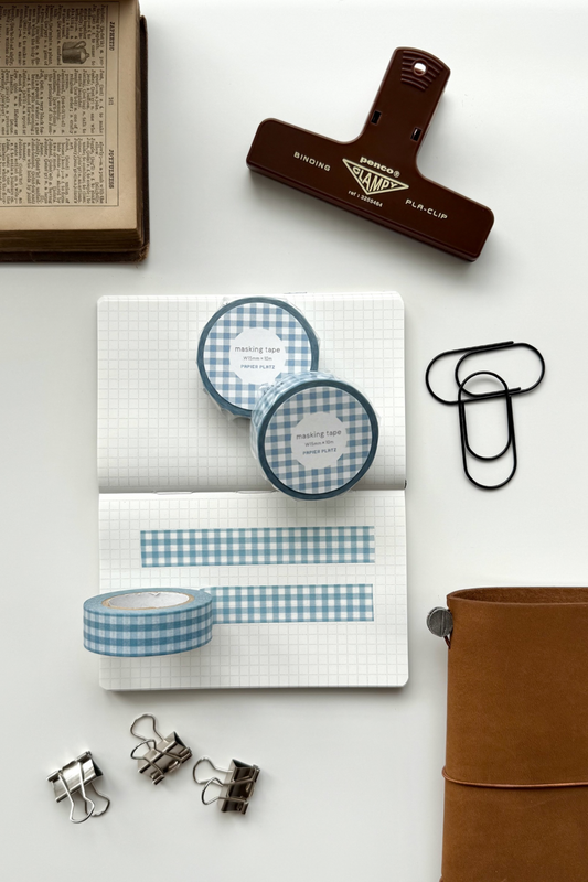 Blue gingham masking tape rolls, grid notebook with tape strips, clips, leather journal, vintage book, and red binding clip in a flat lay stationery setup.