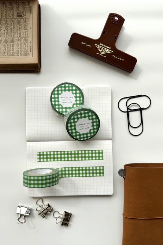 Green gingham masking tape rolls, grid notebook with tape strips, clips, leather journal, vintage book, and red binding clip in a flat lay stationery setup.