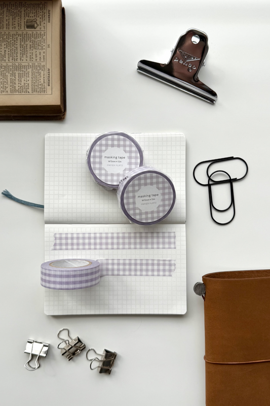 Lavender gingham masking tape rolls, grid notebook with tape strips, clips, leather journal, vintage book, and red binding clip in a flat lay stationery setup.


