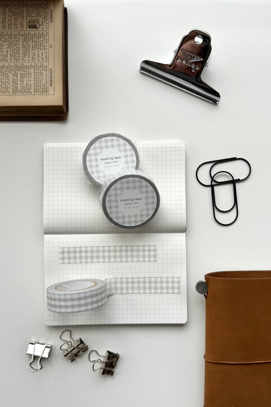 Light grey gingham masking tape rolls, grid notebook with tape strips, clips, leather journal, vintage book, and red binding clip in a flat lay stationery setup.

