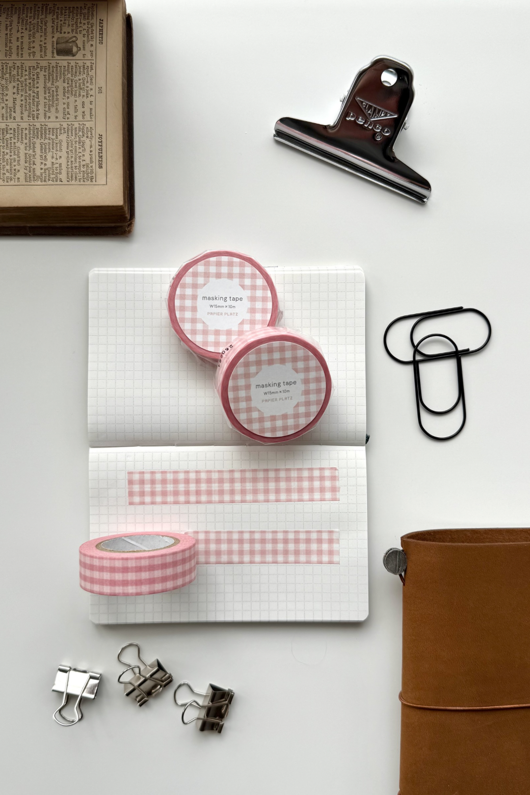 Pink gingham masking tape rolls, grid notebook with tape strips, clips, leather journal, vintage book, and red binding clip in a flat lay stationery setup.