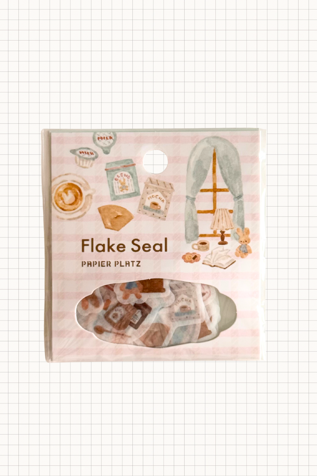 Papier Platz Flake Seal stickers in a soft pink packet, featuring hand-drawn milky drinks, cakes and cosy window scenes with books and lamps on a pale background, shown on grid paper