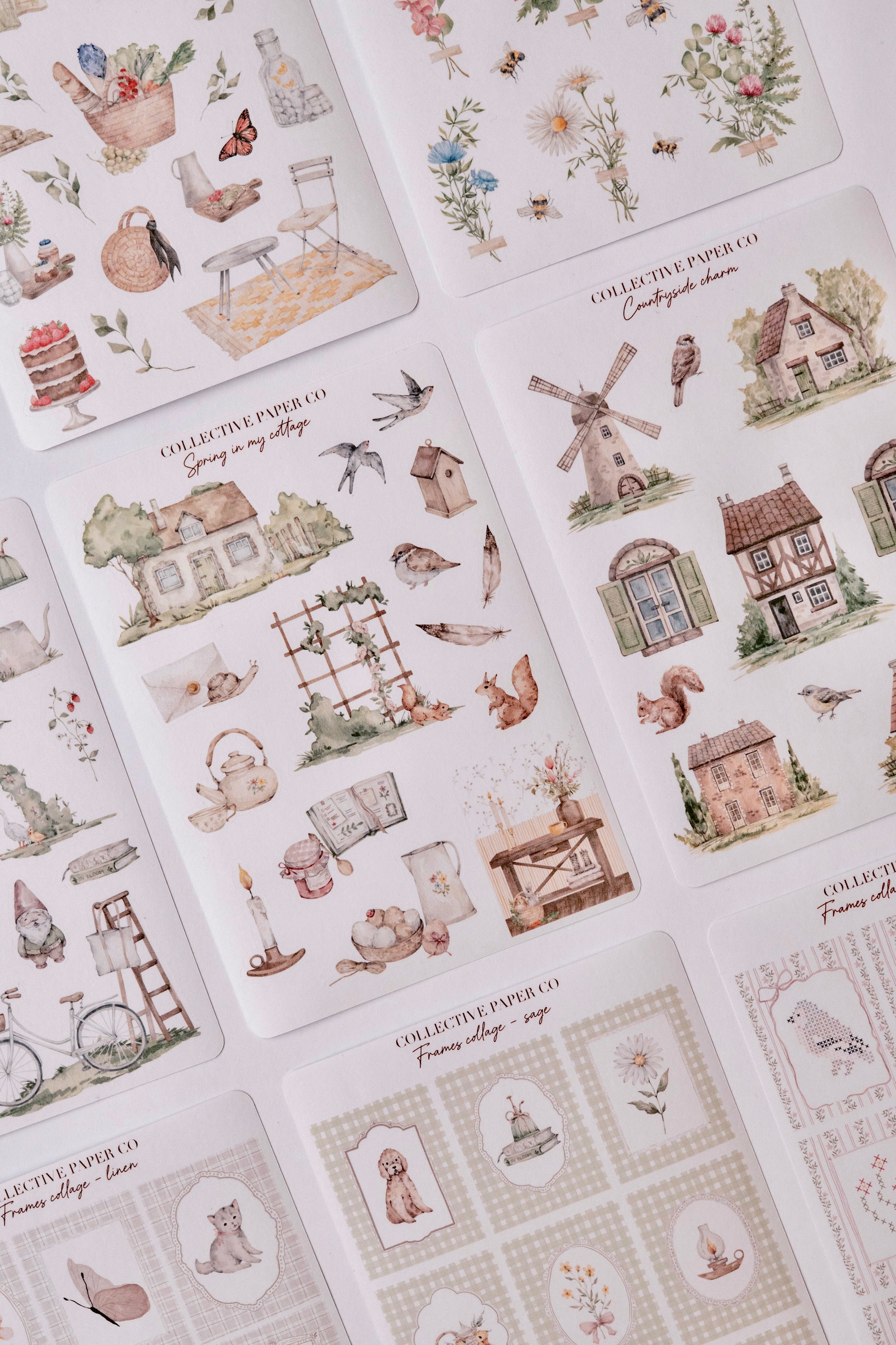 A close-up photo displays an assortment of delicate watercolour-style washi tape sheets from the Colio Paper Co. Spring Village collection, featuring charming motifs like quaint cottages, birds, flowers, squirrels, butterflies, lanterns, teapots, baskets of fruit, and cats on a light background.