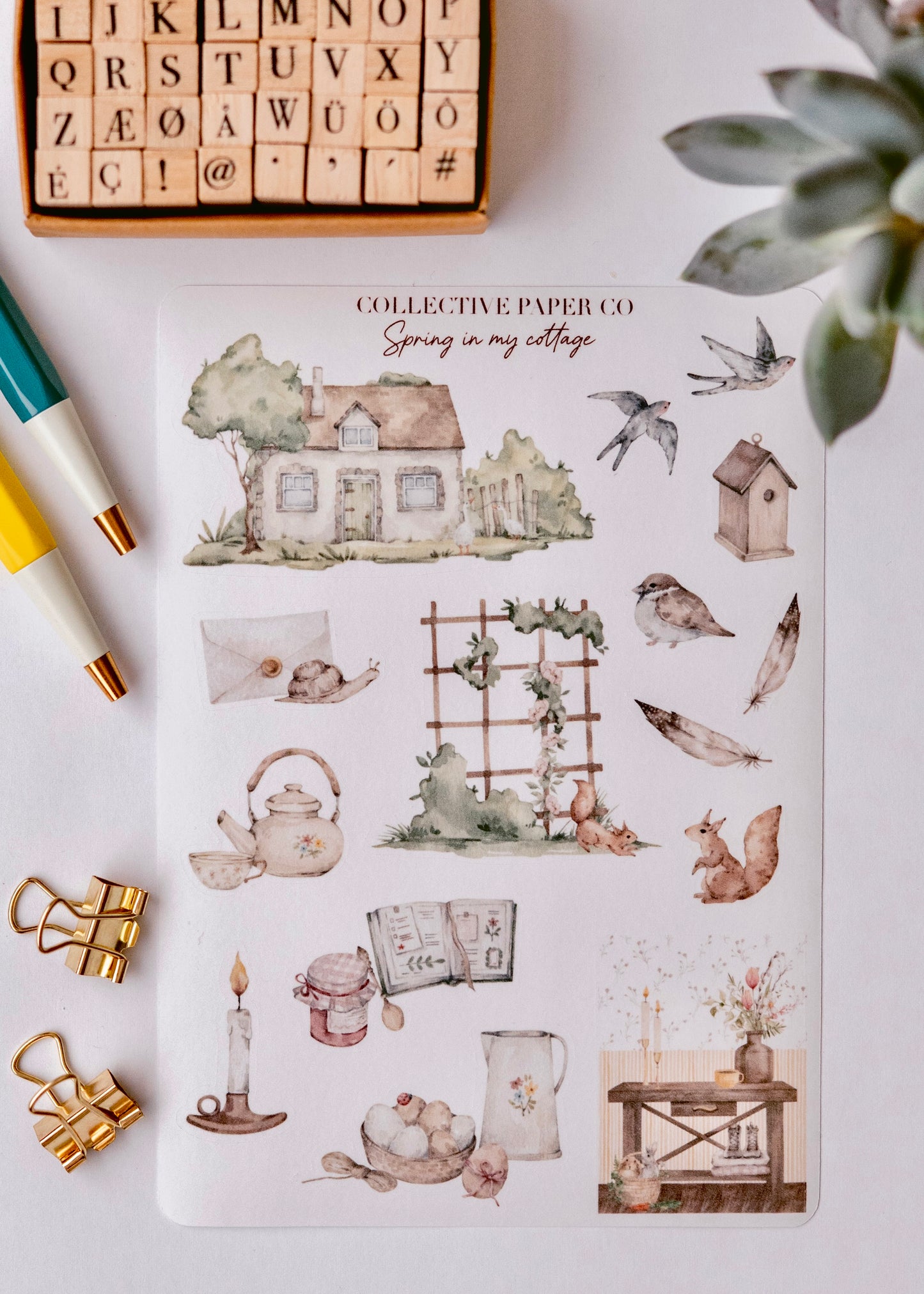 Collective Paper Co.'s Spring in My Village washi tape features watercolor cottages, birds, snails, teapots, and lanterns, styled with stamps, pens, clips, and a succulent.
​

