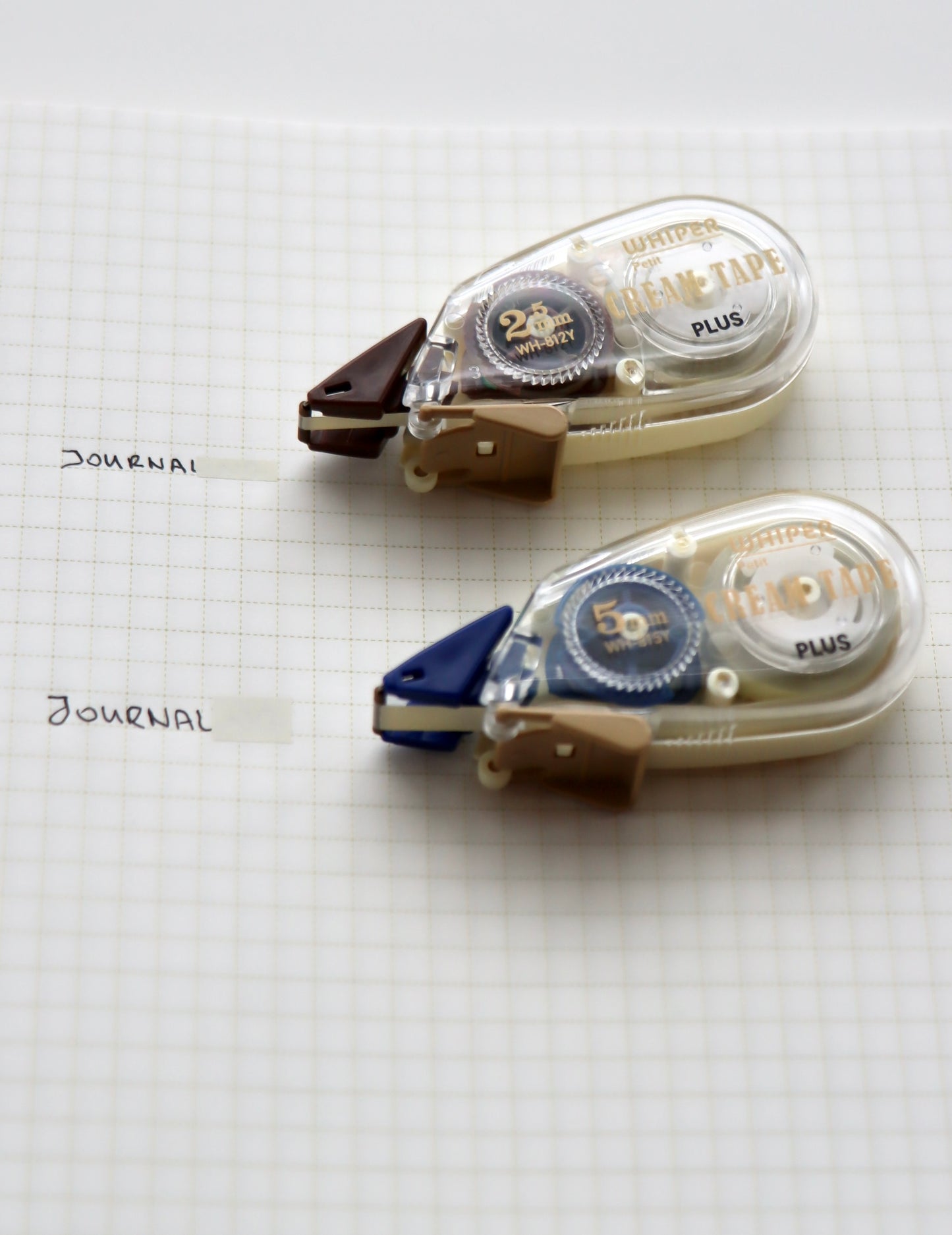 Two Whisper Chara Tape Plus dispensers—one brown for 25mm width (10m) and one blue for 50mm width (9m)—with built-in cutters, arranged on graph paper.
​

