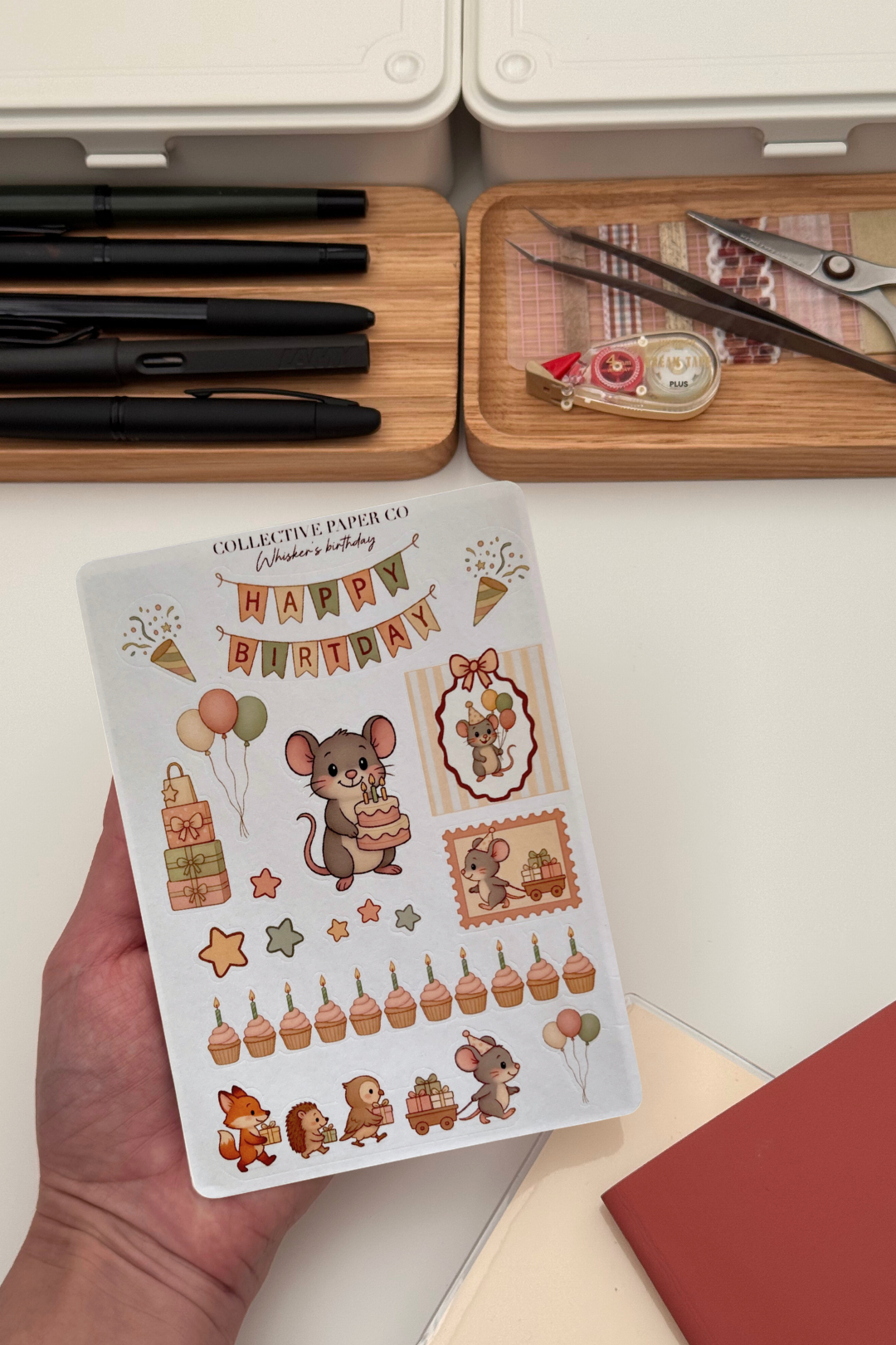Sheet of decorative stickers with birthday-themed designs held by a hand, with stationery items in the background.
