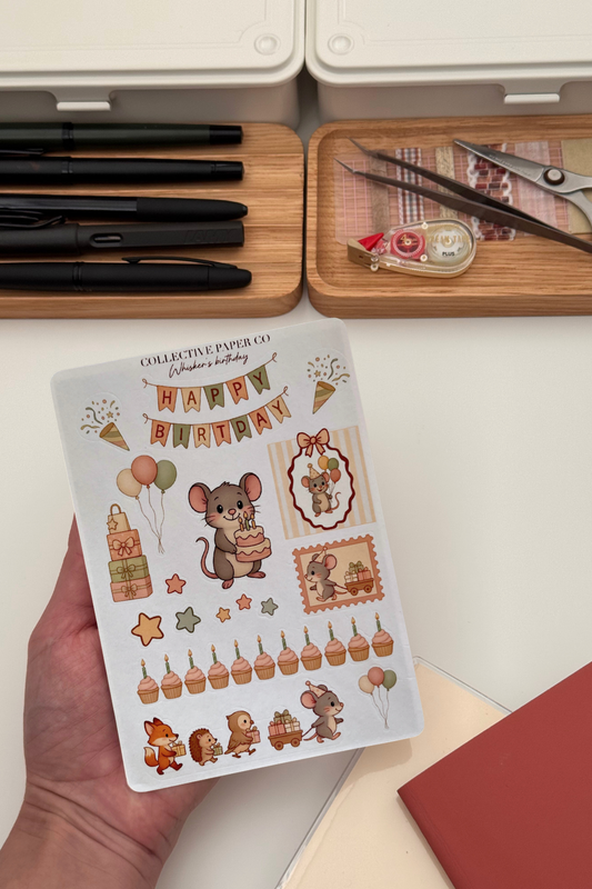 Sheet of decorative stickers with birthday-themed designs held by a hand, with stationery items in the background.