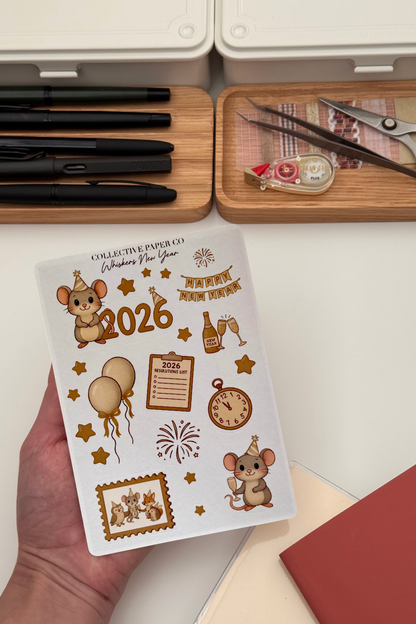 Sheet of decorative stickers with 2026 design held by a hand, with stationery items in the background.