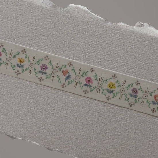 Papier Platz floral masking tape roll featuring a repeating border of tiny multicoloured flowers and leaves on cream tape, shown on grid background with a swatch of the 20 mm wide 5 m tape unrolled