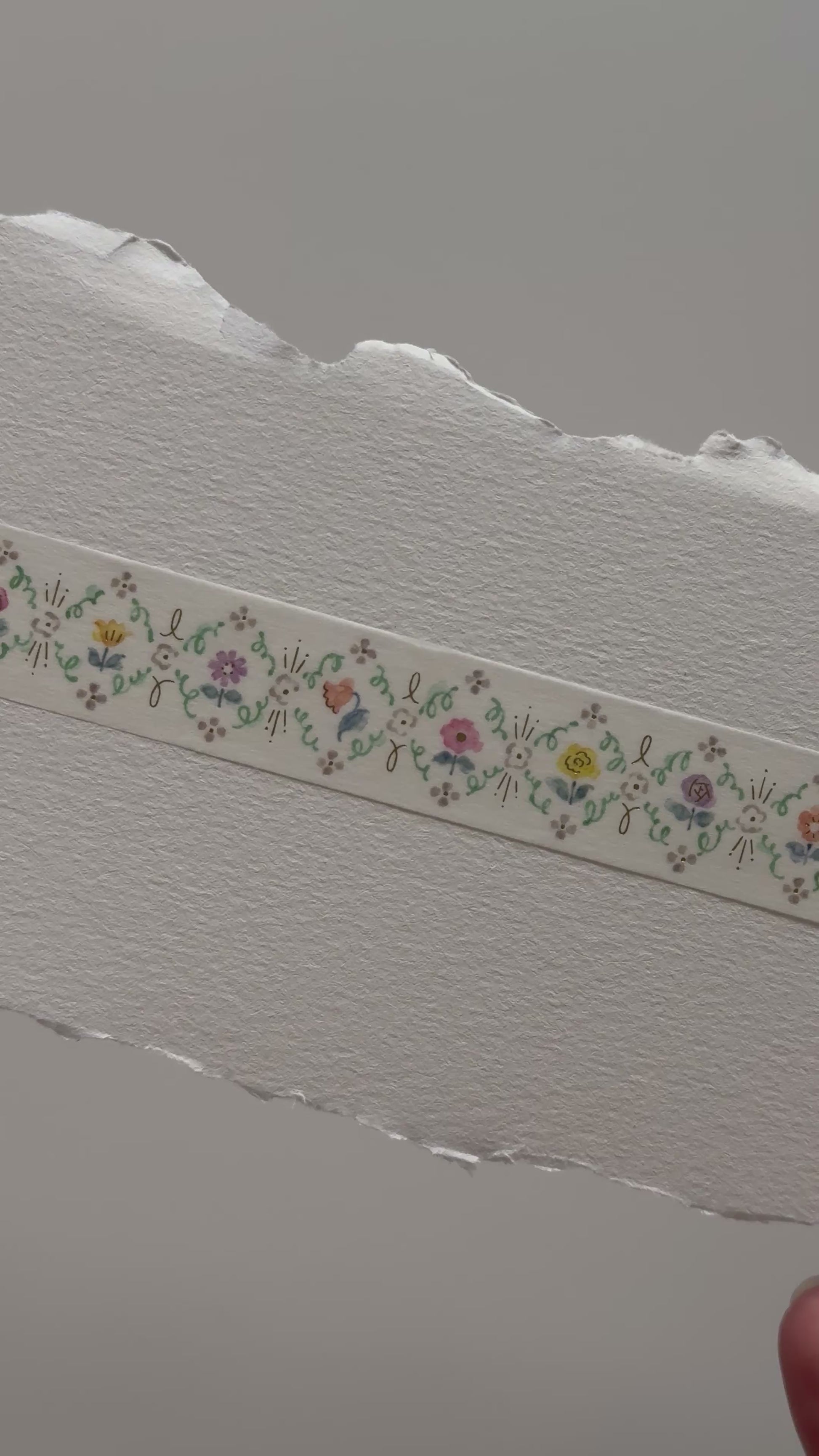 Papier Platz floral masking tape roll featuring a repeating border of tiny multicoloured flowers and leaves on cream tape, shown on grid background with a swatch of the 20 mm wide 5 m tape unrolled